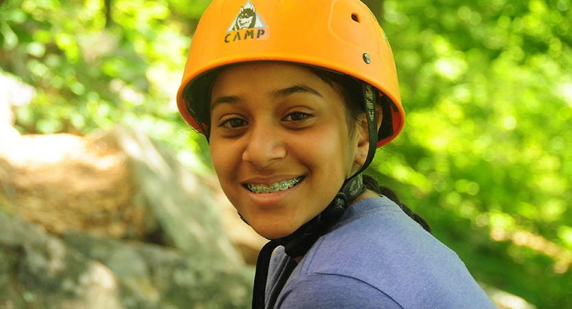 A young person wearing a helmet smiles at the camera. There is blurred greenery in the background. 